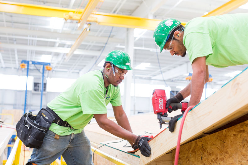 Two workers use power tools to build a manufactured home in a large warehouse. They’re wearing protective equipment and working as a team to ensure quality and safety.